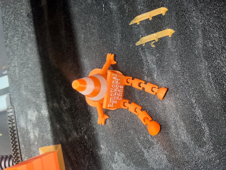 May include: An orange and white traffic cone figure with articulated limbs and a square sign reading Emotional Support Cone. The figure has two arms and two legs with rounded feet. Two separate gold-colored blade-like accessories are included.