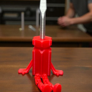 May include: A red, 3D-printed screwdriver with arms and legs, giving it a playful, anthropomorphic appearance. The screwdriver's metal blade is silver. The tool is sitting on a wooden surface.