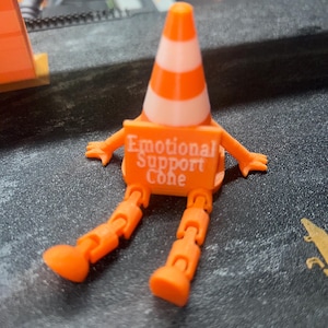 May include: A 3D printed orange and white traffic cone figurine with articulated arms and legs. The cone features a rectangular sign on its front with the white text "Emotional Support Cone" printed on an orange background, sitting in a seated position.