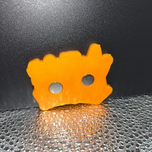 May include: An orange, irregularly shaped magnet with two circular holes. The magnet is made of a solid material and has a textured surface. The magnet is likely used for holding items on a metal surface.