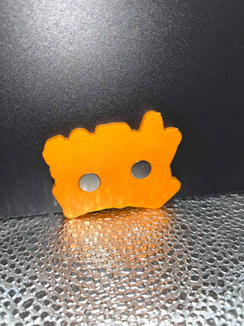 May include: An orange, irregularly shaped magnet with two circular holes. The magnet is made of a solid material and has a textured surface. The magnet is likely used for holding items on a metal surface.