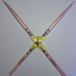Pode incluir: Duas baquetas de bateria de madeira cruzadas em forma de X, presas por um suporte de plástico amarelo. As baquetas são castanhas claras com texto e gráficos pretos. O texto "AMERICAN CLASSIC EXTREME 5A" é visível.