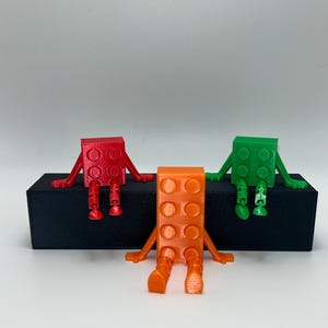 Könnte beinhalten: Drei farbenfrohe, 3D-gedruckte Blockfiguren mit Armen und Beinen sind auf einer schwarzen rechteckigen Basis angeordnet. Die Figuren sind rot, orange und grün, jede mit einem einzigartigen Design. Die Figuren sitzen mit baumelnden Beinen.