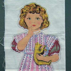 May include: A fabric cutout of a young girl in a pink dress with a white and blue pattern, holding a yellow bear. The girl has blonde hair and is wearing pink socks and yellow shoes. The image is on a white background.