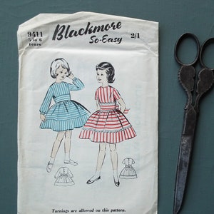 Op de afbeelding: Een vintage naaipatroon voor een meisjesjurk. Het patroon is voor maten 5 tot 6 jaar oud en is van de Blackmore So-Easy-lijn. Het patroon toont twee meisjes die jurken dragen, een in blauw-witte strepen en de andere in rood-witte strepen. Het patroon is gemarkeerd met 9411.