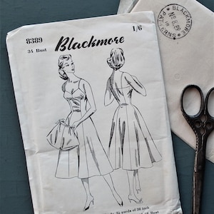 May include: Vintage sewing pattern titled "Blackmore" featuring a dress design with a fitted bodice and flared skirt. The pattern includes illustrations of the dress, measurements, and additional sizes. A pair of vintage scissors and a stamped envelope are also visible.