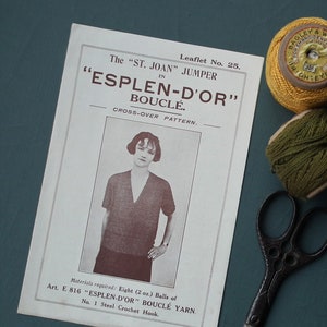 May include: A vintage knitting pattern leaflet for "The St. Joan Jumper" in "Esplen-D'Or" Bouclé. The leaflet has a black and white image of a woman wearing the jumper. There are also two spools of thread and a pair of scissors.