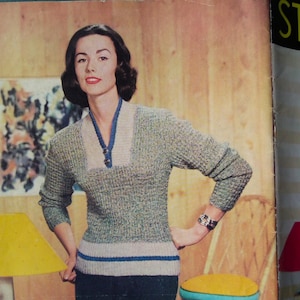 May include: A woman wearing a gray and blue knitted sweater with a white collar and a blue stripe at the waist. She is standing in a room with a yellow lamp and a chair with a yellow cushion.