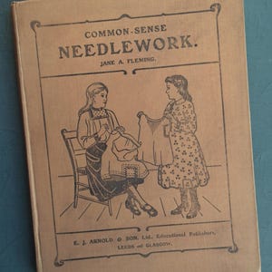 May include: A vintage book titled "COMMON-SENSE NEEDLEWORK" by Jane A. Fleming. The cover features a black and white illustration of two women, one seated and sewing, the other holding up a garment. Published by E. J. Arnold & Son, Ltd.