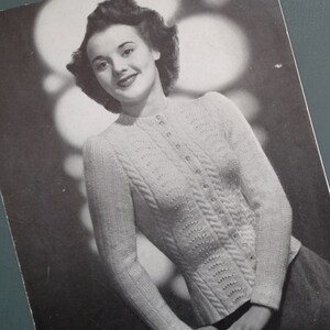 May include: A black and white photo of a woman wearing a knitted cardigan with a cable knit pattern and button closure.