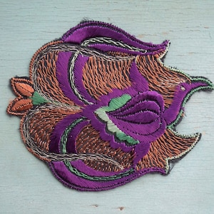 May include: A purple, orange, and green embroidered patch in the shape of a stylized flower. The patch is made of satin and has a detailed design.