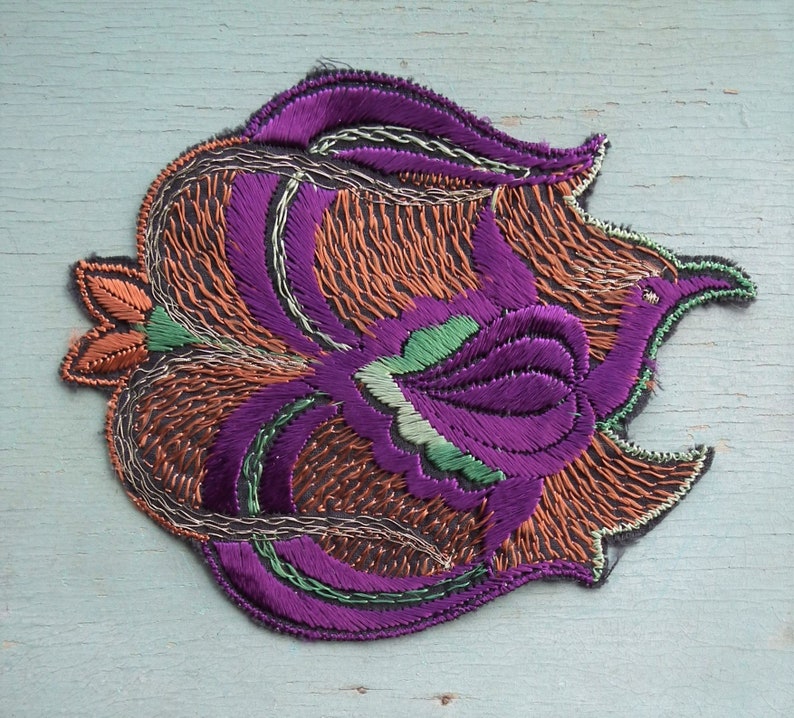 May include: A purple, orange, and green embroidered patch in the shape of a stylized flower. The patch is made of satin and has a detailed design.
