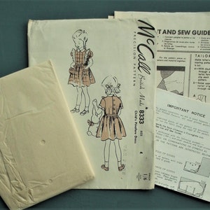 Op de afbeelding: Vintage McCall's naaipatroon voor een kinder pinafore jurk. Het patroonpakket, met het label "British Made", toont illustraties van de jurk en bevat instructies en een knipgids. Maatinformatie is ook zichtbaar.