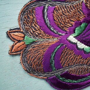 May include: A close-up of a purple, green, and orange embroidered floral design. The embroidery is done on a dark fabric and has a detailed, intricate pattern.