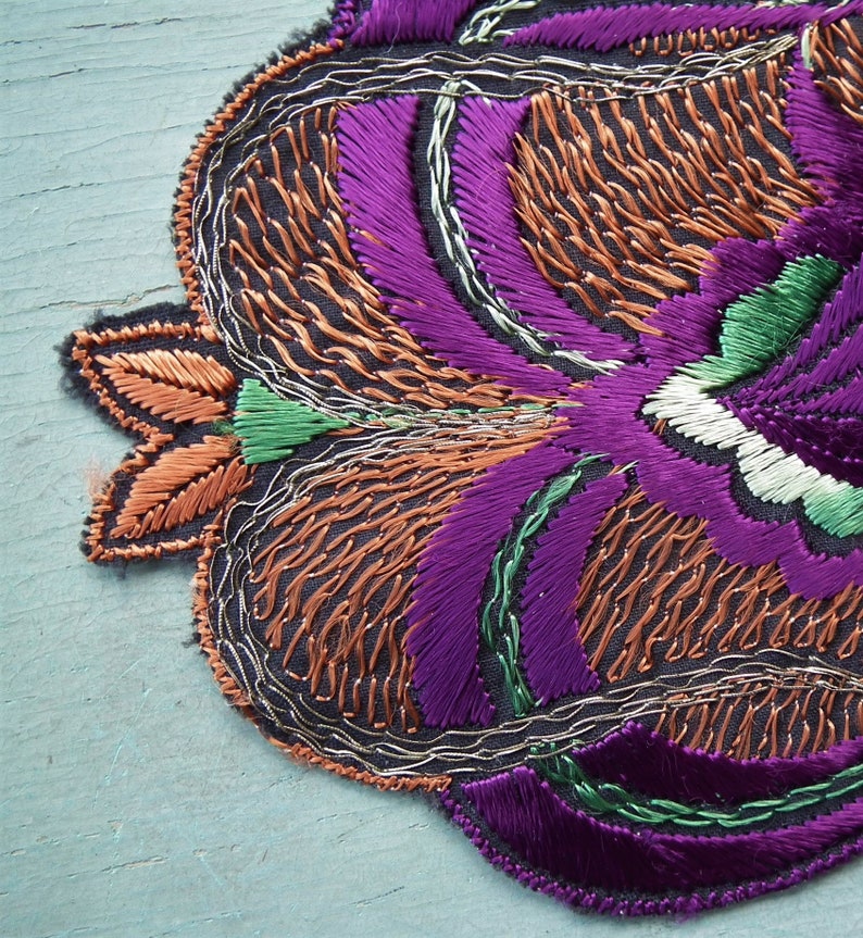 May include: A close-up of a purple, green, and orange embroidered floral design. The embroidery is done on a dark fabric and has a detailed, intricate pattern.