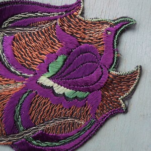 May include: A purple, green, and orange embroidered patch with a floral design. The patch is made of fabric and has a scalloped edge.
