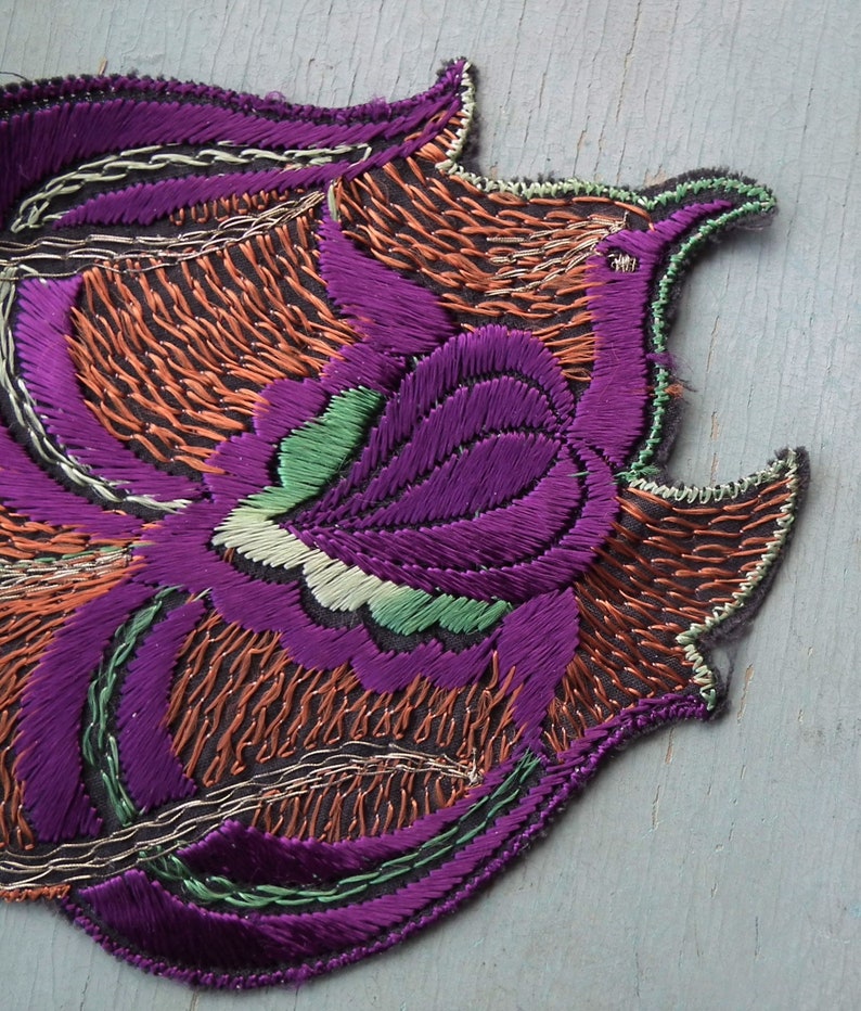 May include: A purple, green, and orange embroidered patch with a floral design. The patch is made of fabric and has a scalloped edge.