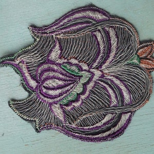 May include: A black embroidered patch with a purple, white, and green floral design. The patch has a scalloped edge and is perfect for adding a unique touch to clothing or accessories.