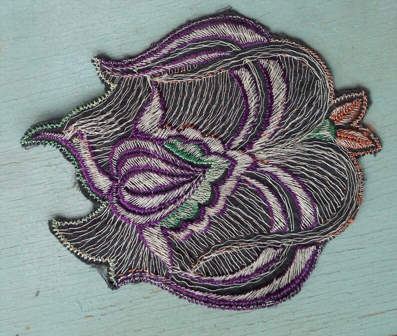 May include: A black embroidered patch with a purple, white, and green floral design. The patch has a scalloped edge and is perfect for adding a unique touch to clothing or accessories.