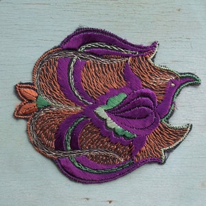 May include: A purple, green, and orange embroidered patch with a floral design. The patch is made of fabric and has a textured surface.