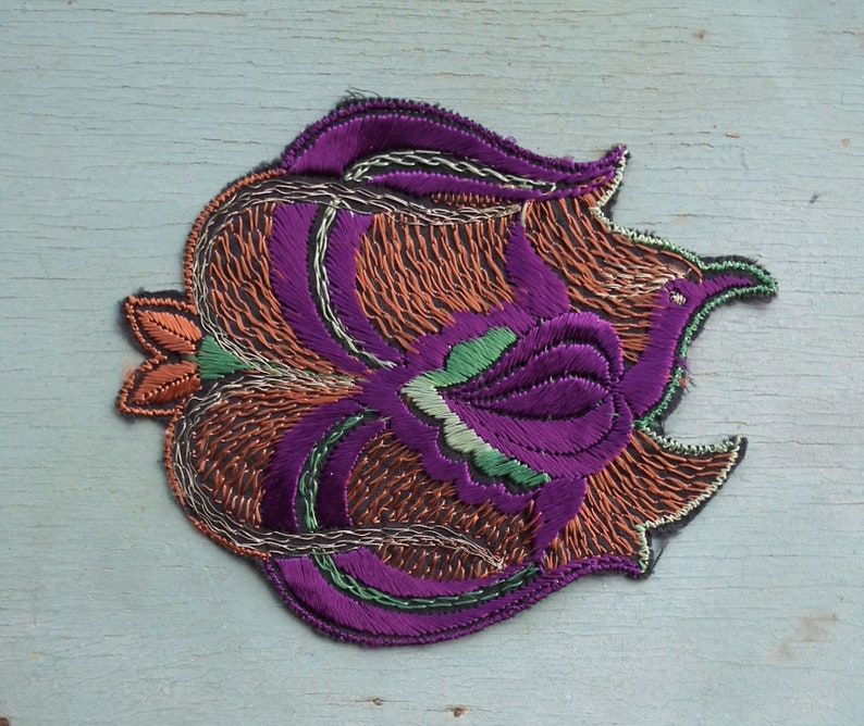 May include: A purple, green, and orange embroidered patch with a floral design. The patch is made of fabric and has a textured surface.