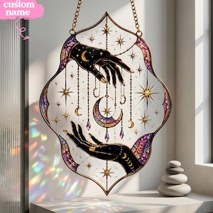 Peut inclure: Un attrape-soleil en vitrail de forme ovale pointue, représentant deux mains noires tendant vers un croissant de lune central. Le motif inclut des étoiles dorées, des chaînes pendantes et des touches violettes. Le texte custom name est visible en haut à gauche.