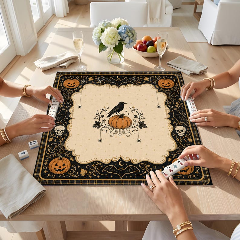 May include: A square game mat featuring a cream-colored center with a black crow perched on a pumpkin, surrounded by a black border decorated with orange jack-o'-lanterns, skulls, bats, and spiders. The mat is placed on a wooden table.