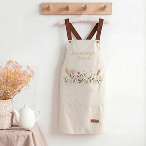 Op de afbeelding: Crèmekleurig schort met bruine bandjes, hangend aan een houten rek. Het schort heeft de tekst "Sourdough Queen" en een geborduurd bloemendesign. Een zak is zichtbaar aan de voorkant. Het schort hangt aan een witte hanger.