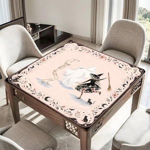 May include: A square table cover featuring a pale pink background with a central illustration of a skeleton, a white ghost, and a witch holding a broom. The border is decorated with black floral vines, crescent moons, bats, and small black cats.