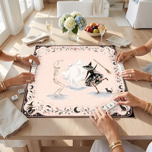 May include: A square mahjong mat featuring a pale pink center with illustrations of a skeleton, a ghost, and a witch with a broom and black cat. The border is decorated with black floral patterns, moons, and stars, with mahjong tiles placed around.