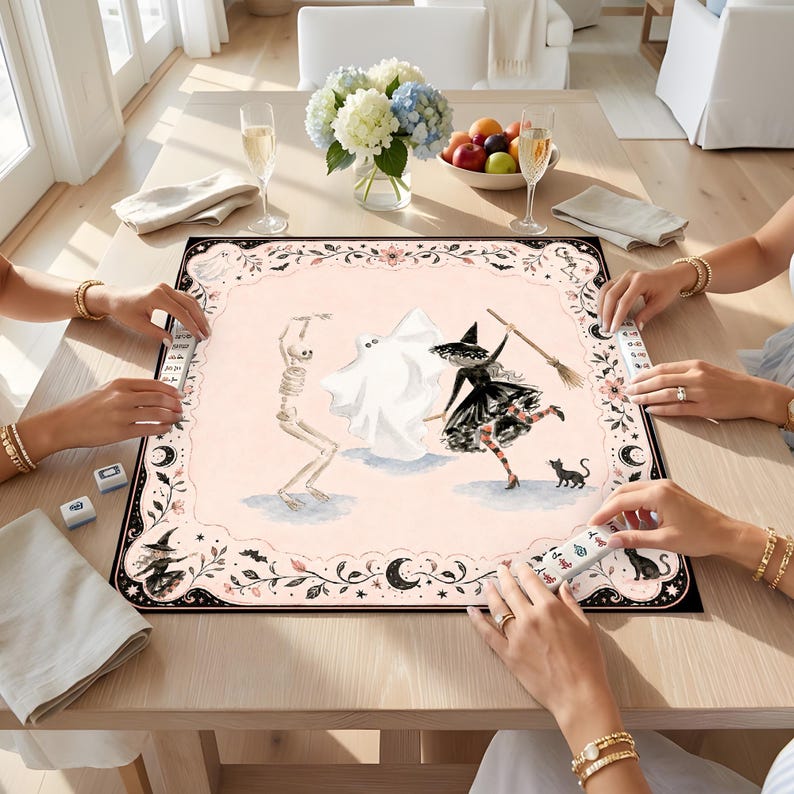 May include: A square mahjong mat featuring a pale pink center with illustrations of a skeleton, a ghost, and a witch with a broom and black cat. The border is decorated with black floral patterns, moons, and stars, with mahjong tiles placed around.