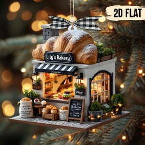 May include: A miniature bakery ornament featuring a croissant dusted with powdered sugar. The bakery has a black and white striped awning, a sign that reads "Lily's Bakery", and a chalkboard sign. The ornament is decorated with miniature baked goods and greenery.