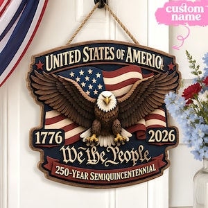 May include: A wooden sign with a patriotic theme, featuring an American flag and a bald eagle. The sign displays the text "United States of America", "1776", "2026", and "We the People". It is designed for hanging, with a rope attached.