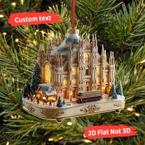 Puede incluir: Adorno navideño que representa una réplica detallada de una catedral. El adorno tiene una base crema con detalles dorados, un tranvía rojo y pequeños árboles verdes. El texto "Custom text" es visible. Incluye una cuerda roja.