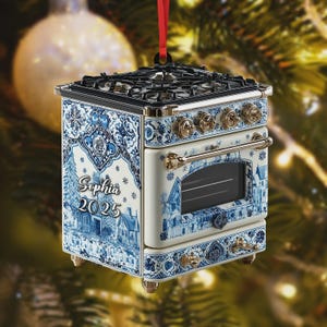 以下が含まれることがあります： 青と白のデルフトデザインのミニチュアストーブのクリスマスオーナメント。オーナメントは黒いコンロ、金色のノブ、赤いリボンが付いています。側面には「Sophia 2025」の文字が見えます。