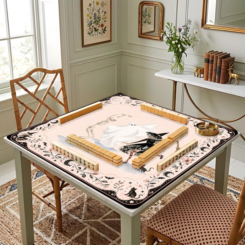May include: A square mahjong table mat featuring a pale pink surface with illustrations of a skeleton, a ghost, and a witch. The border is decorated with black floral patterns, moons, and cats. Tan mahjong tiles are arranged on the mat.
