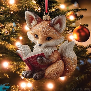 Peut inclure: Une décoration de sapin de Noël en forme de renard au pelage orange et blanc, tenant un livre rouge ouvert. Le renard a des pattes et des oreilles brun foncé, avec des reflets scintillants sur sa fourrure. Une boucle en ruban rouge est fixée.