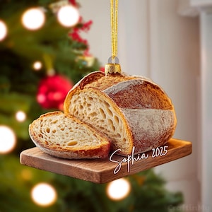 Puede incluir: Un adorno navideño colgante con forma de hogaza de pan de masa madre sobre una pequeña tabla de madera. El pan tiene una corteza dorada y un interior aireado. El texto Sophia 2025 está escrito en blanco en la tabla. Incluye un cordón dorado.