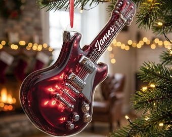 May include: A glossy, deep red electric guitar ornament featuring silver hardware details and the name James printed in white script on the neck. The ornament hangs from a thin red ribbon, designed as a decorative piece for a holiday tree.