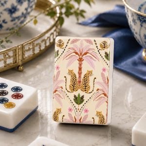 Puede incluir: Una ficha de mahjong rectangular con fondo color crema y un patrón repetitivo de dos leopardos frente a frente bajo una palmera rosa, acentuado con pequeñas flores rosas y follaje verde, con un acabado liso y brillante.