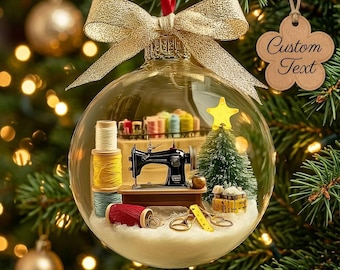 Puede incluir: Un adorno esférico de cristal transparente que contiene una escena de costura en miniatura con una máquina de coser negra, carretes de hilo de colores, un pequeño árbol de Navidad verde con una estrella amarilla y tijeras doradas, rematado con un lazo dorado y una etiqueta de madera.