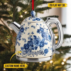 Puede incluir: Un adorno de tetera crema con un estampado floral azul y un asa y boquilla plateadas. El adorno tiene el nombre "Jennifer" en cursiva y el texto "CUSTOM NAME HERE". Se adjunta una cinta roja para colgar.