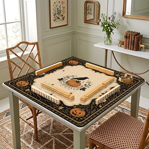 May include: A square mahjong mat featuring a cream-colored center with a black crow, a pumpkin, and spiderweb illustrations. The border is black with orange jack-o'-lanterns and intricate scrollwork. Four sets of wooden mahjong tiles are arranged.
