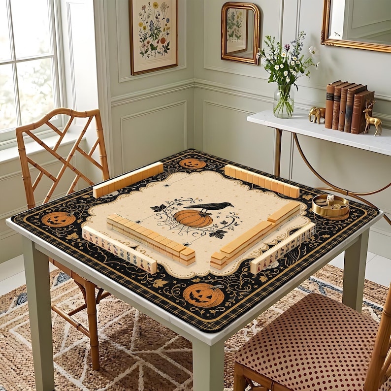 May include: A square mahjong mat featuring a cream-colored center with a black crow, a pumpkin, and spiderweb illustrations. The border is black with orange jack-o'-lanterns and intricate scrollwork. Four sets of wooden mahjong tiles are arranged.