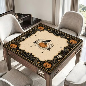 May include: A square tablecloth featuring a cream-colored center with a black crow perched on a pumpkin and spiderweb design. The border is black with orange jack-o'-lanterns, white skulls, spiderwebs, and leafy vine patterns.