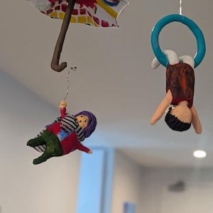 May include: A whimsical mobile featuring three handmade figures. One figure is suspended from a colorful umbrella, another hangs from a blue hoop, and the third is a figure with a scarf. The figures are suspended from the ceiling.