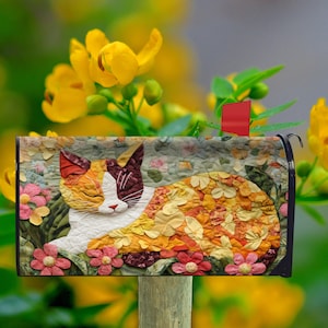 May include: A decorative mailbox cover featuring a colorful, quilted design of a cat resting among flowers. The cat has orange, white, and brown fur. Pink and yellow flowers surround the cat on a green background. A red flag is raised.