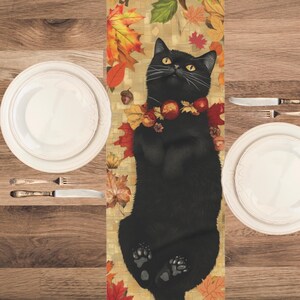 May include: A table runner featuring a black cat illustration surrounded by autumn leaves and apples. Two white plates, cutlery, and a wooden table create a fall theme. The cat is lying on its back, gazing upwards.