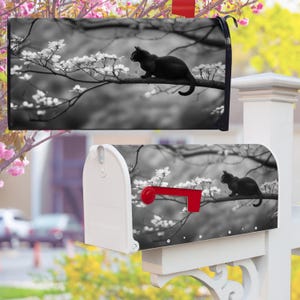 Könnte beinhalten: Schwarz-weißer Briefkasten mit einer schwarzen Katze auf einem Ast mit weißen Blüten. Der Briefkasten ist weiß mit einer roten Fahne.