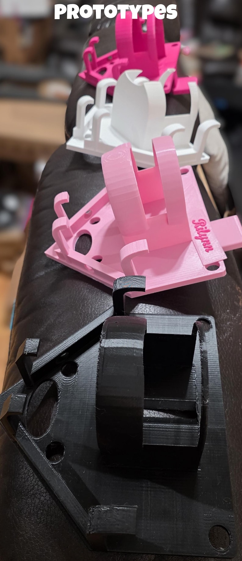 May include: A collection of 3D-printed prototypes in various stages of completion. The prototypes are in pink, white, and black. The word "PROTOTYPES" is printed at the top of the image. One pink prototype has the name "Rilynn" printed on it.